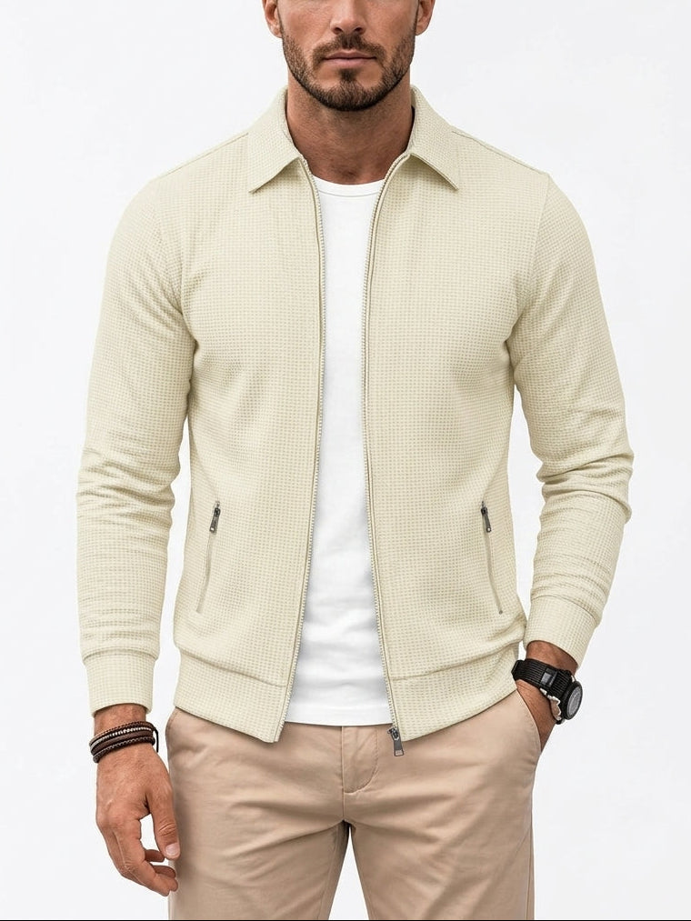 A man wearing a light beige textured zip-up jacket over a white t-shirt and beige trousers, standing against a plain white background.