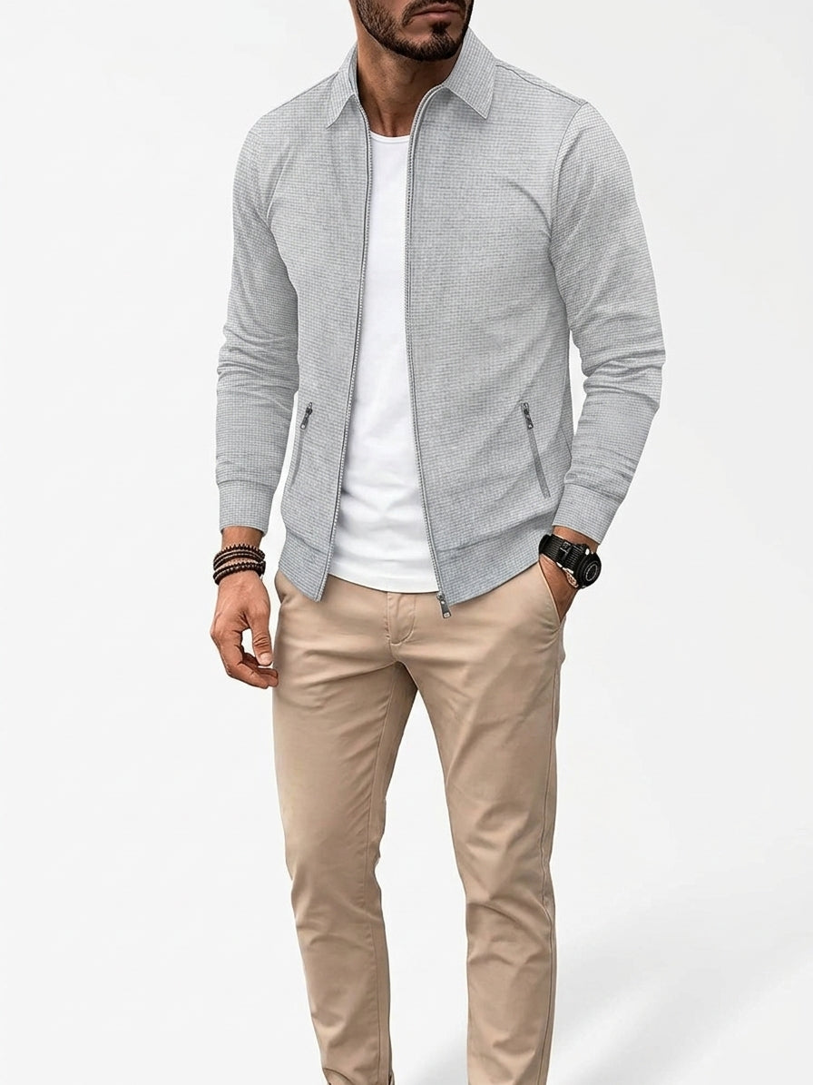 A man wearing a light gray zip-up collared jacket over a white t-shirt and beige pants, standing against a light gray studio background.