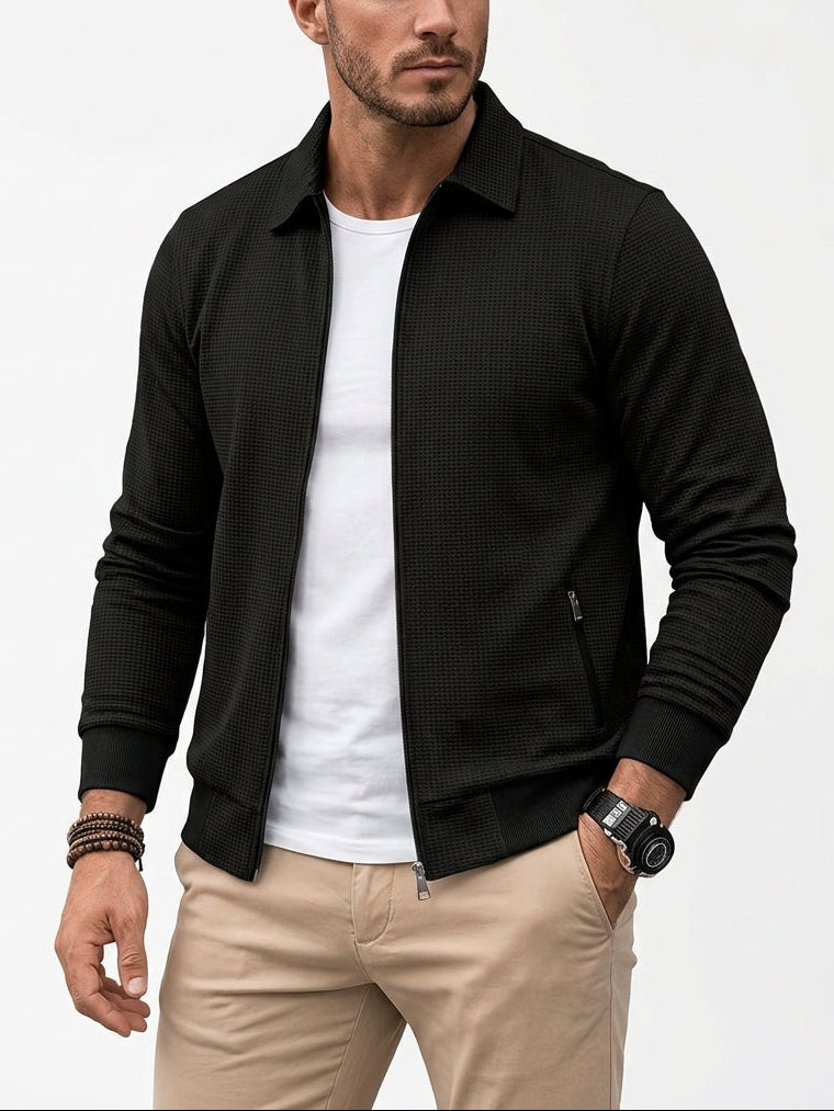 A man wearing a black textured zip-up jacket over a white t-shirt and beige pants, photographed against a clean white studio background.