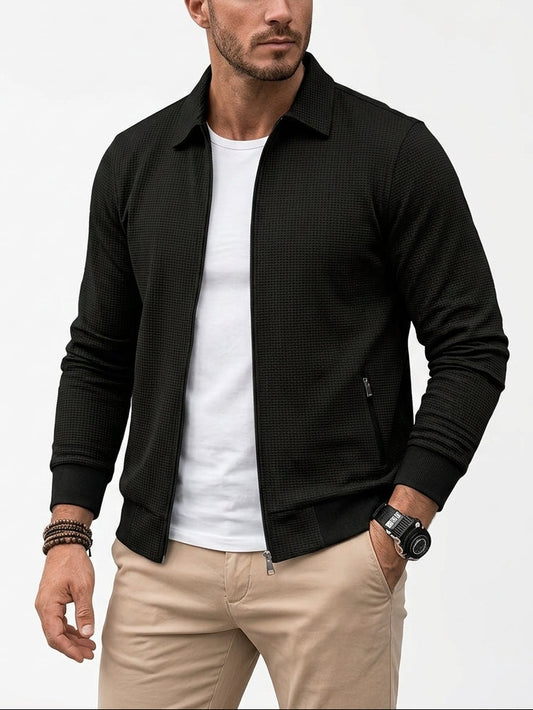 A man wearing a black textured zip-up jacket over a white t-shirt and beige pants, photographed against a clean white studio background.