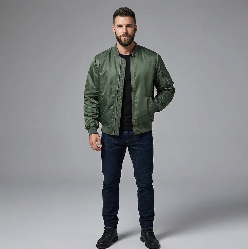 A man wearing an olive green satin bomber jacket over a black t-shirt, dark blue jeans, and black boots, standing against a neutral gray studio background.