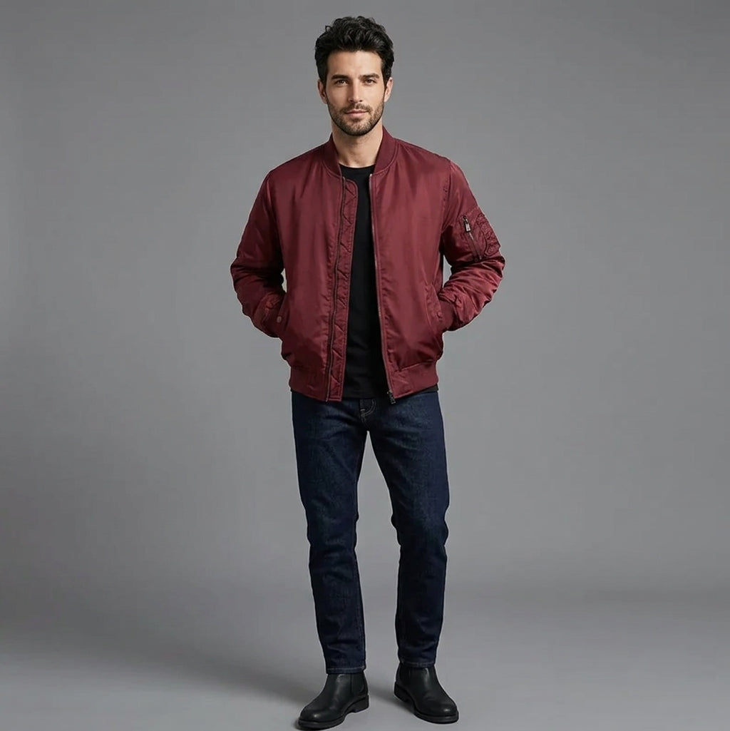 A man modeling a burgundy bomber jacket, dark blue jeans, and black boots against a solid gray studio background.