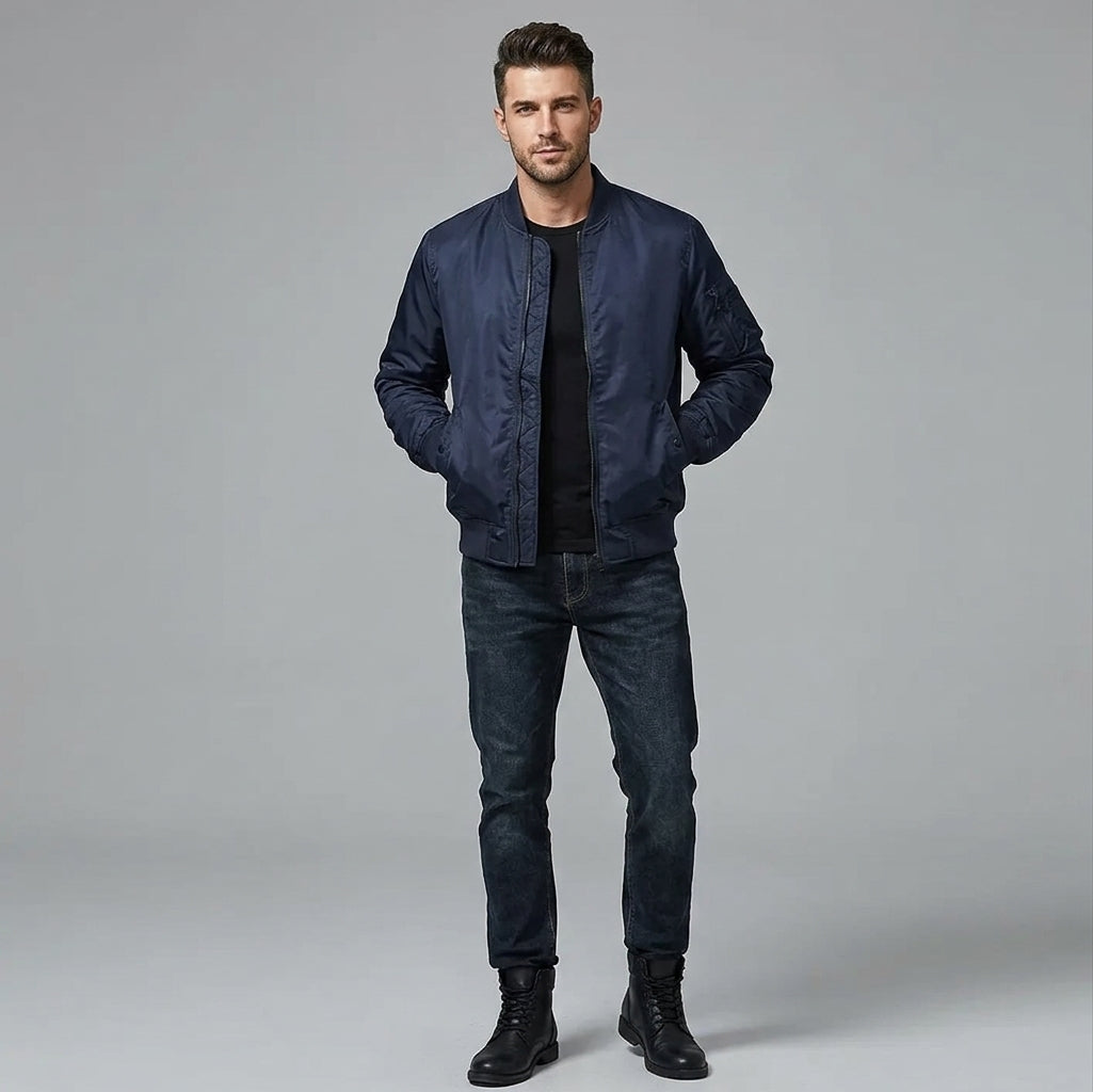 A man modeling a dark navy blue bomber jacket over a black t-shirt, dark blue jeans, and black boots, standing against a solid gray studio background.