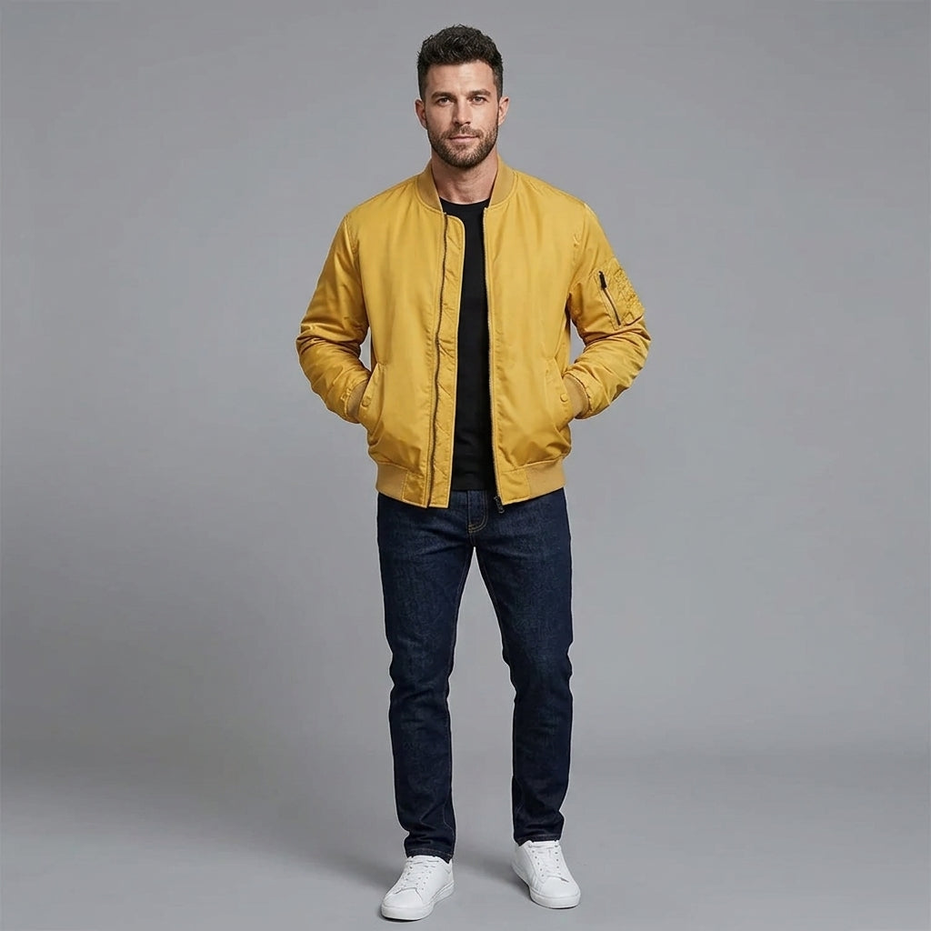 A man modeling a mustard yellow bomber jacket over a black t-shirt, dark blue jeans, and white sneakers, standing against a solid gray studio background.