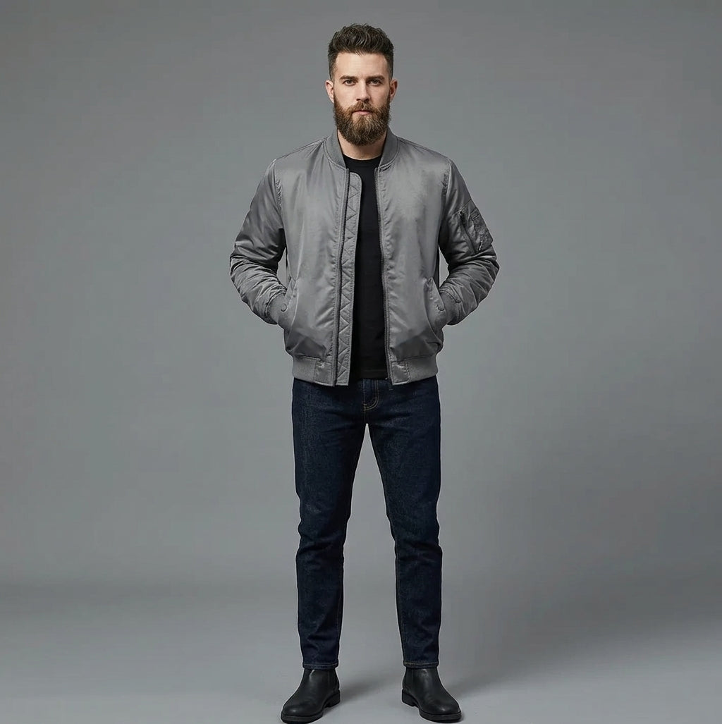 A man with a beard stands against a gray studio background wearing a gray satin bomber jacket over a black t-shirt, dark blue jeans, and black boots.
