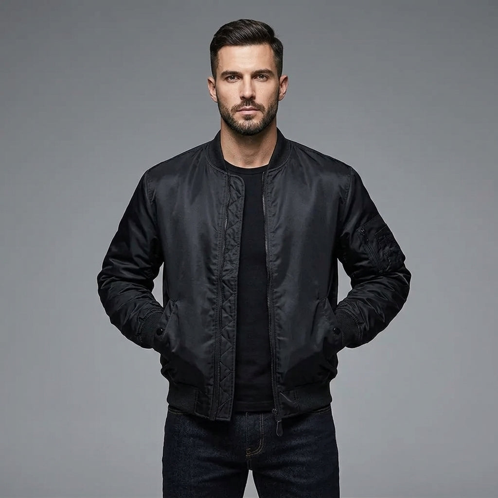 A man wearing a black bomber jacket over a black t-shirt and dark jeans, standing against a solid gray studio background.