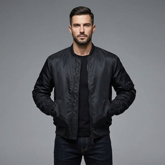 A man wearing a black bomber jacket over a black t-shirt and dark jeans, standing against a solid gray studio background.