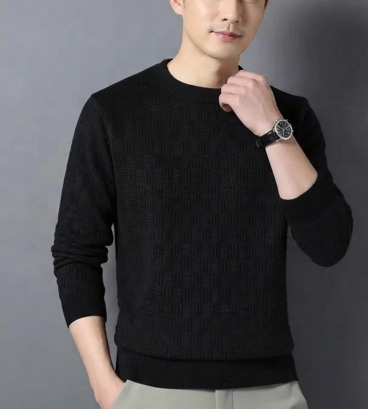 A man wearing a black long-sleeve knit sweater and light gray trousers, standing against a plain gray background. A watch is visible on his wrist