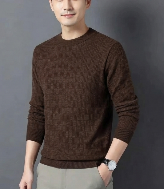 A man wearing a brown ribbed knit crewneck sweater and light gray trousers, standing against a dark gray studio background.
