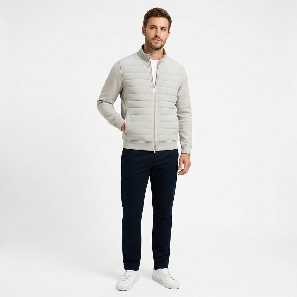 A man modeling a light gray zip-up puffer jacket over a white t-shirt, paired with dark navy pants and white sneakers, against a plain white studio background.
