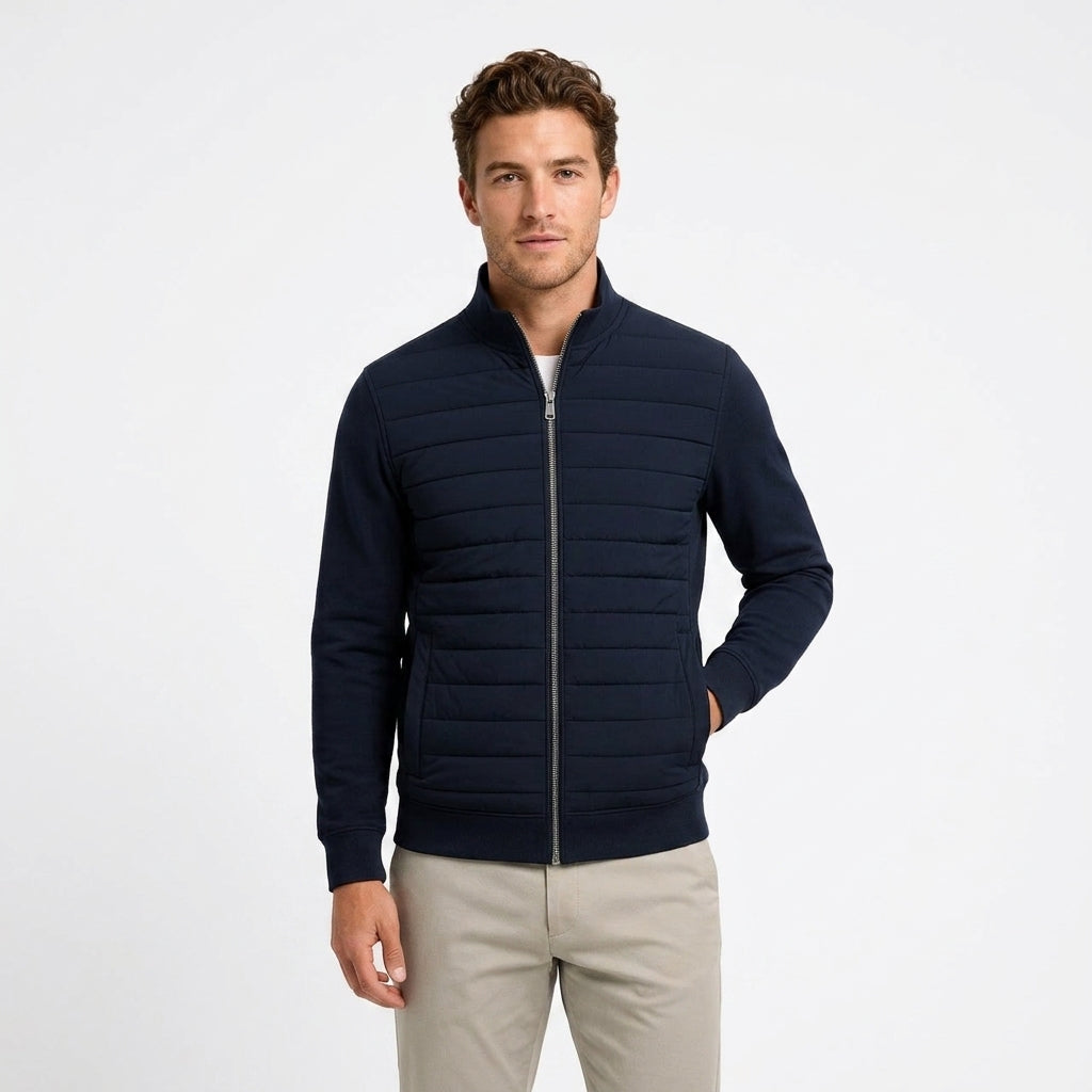 A man wearing a navy blue zip-up quilted jacket and light beige pants, standing against a plain white studio background.