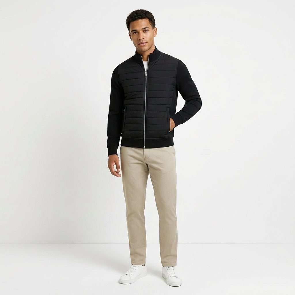 A man modeling a black quilted zip-up jacket with long sleeves, paired with beige trousers and white sneakers, standing against a plain white studio background.