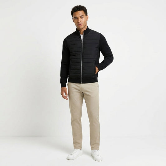 A man modeling a black quilted zip-up jacket with long sleeves, paired with beige trousers and white sneakers, standing against a plain white studio background.