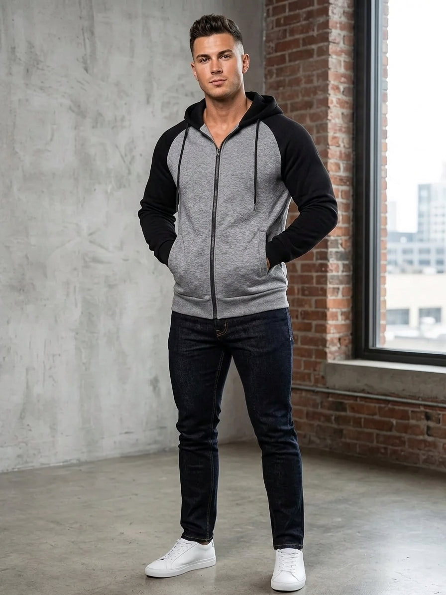 A man wearing a gray and black raglan zip-up hoodie, dark blue jeans, and white sneakers, standing in a loft-style room with a concrete wall, brick column, and large window.