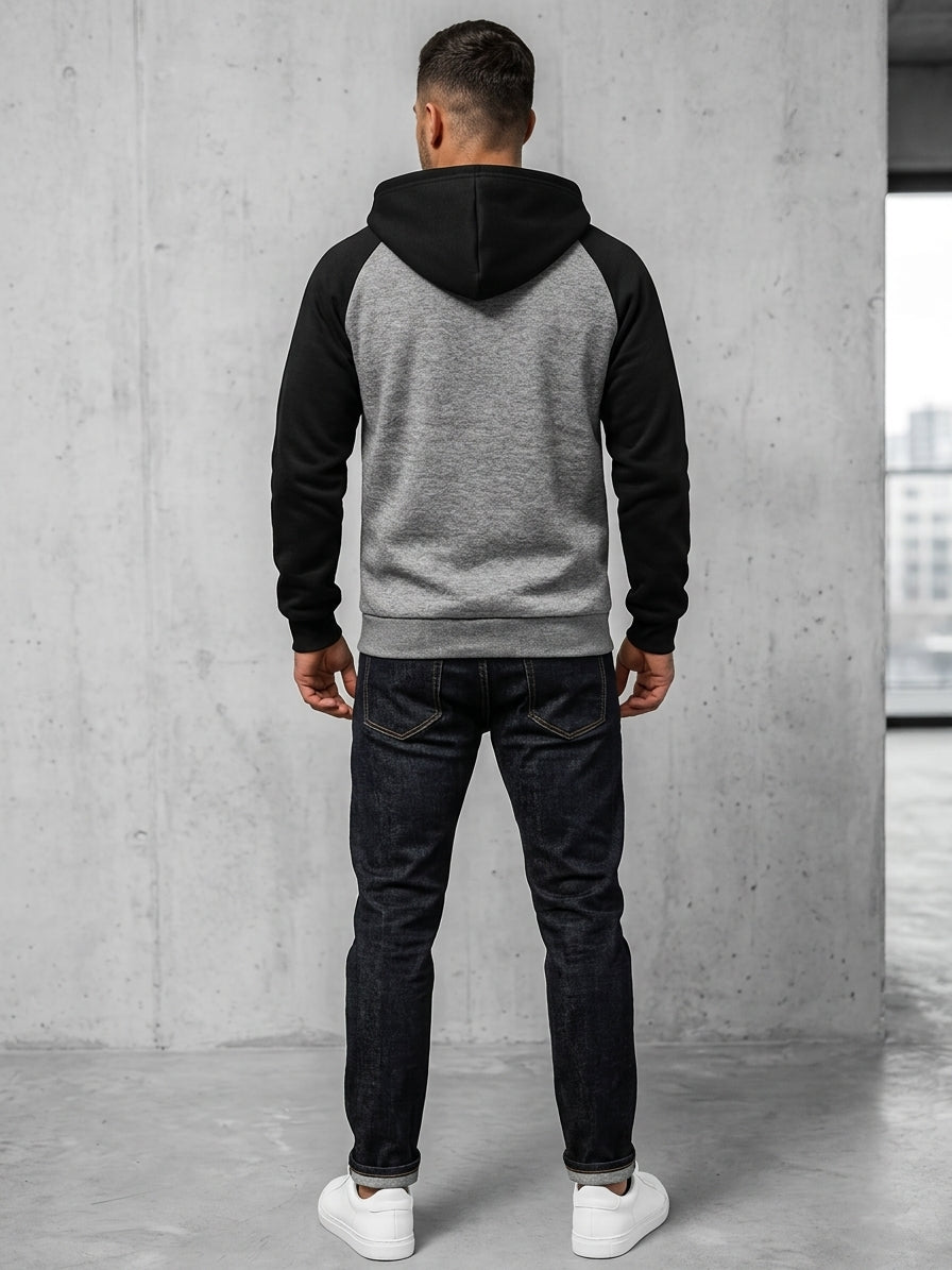 A man viewed from behind wearing a two-tone raglan hoodie with a black hood and sleeves, a heather gray back, and dark wash jeans, standing in a minimalist concrete room.