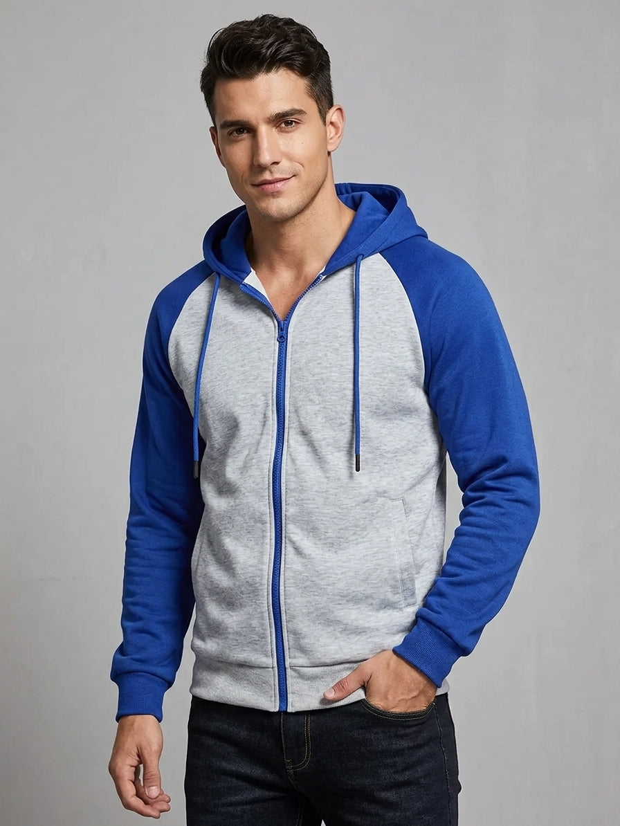 A man wearing a two-tone raglan zip-up hoodie with a heather gray body and royal blue sleeves, hood, and zipper trim, paired with dark jeans against a plain gray studio background.