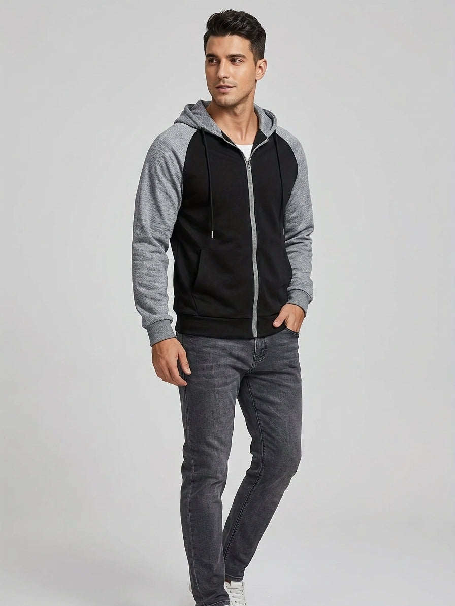 A man modeling a color-blocked zip-up hoodie with gray sleeves and hood, and a black body, paired with dark gray jeans against a light gray studio background.