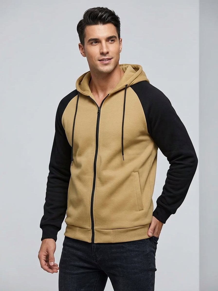 A man wearing a two-tone zip-up hoodie with a tan body and black raglan sleeves, paired with dark jeans, standing against a light gray studio background.
