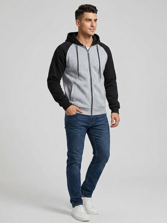A man modeling a color-blocked zip-up hoodie with a heather gray body and black raglan sleeves, paired with blue jeans and white sneakers, against a light gray studio background.
