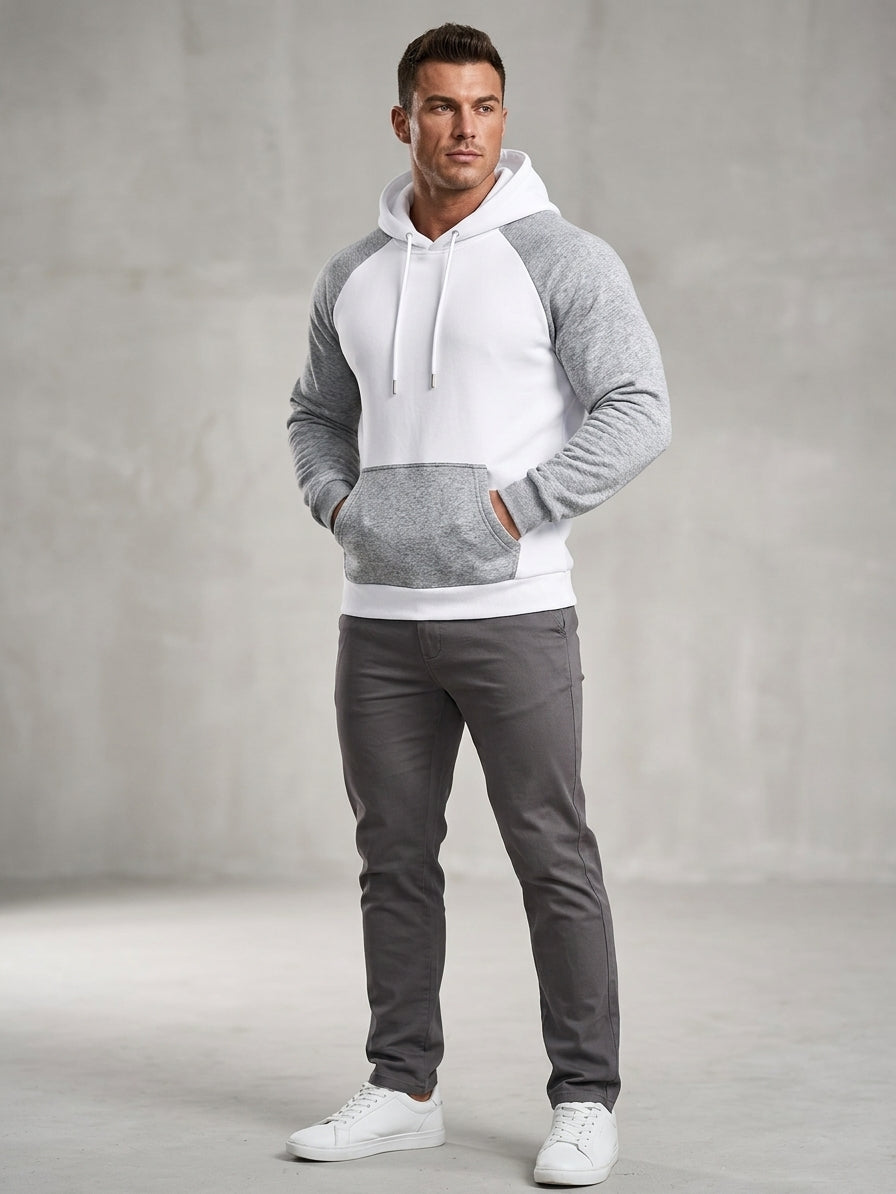 A man modeling a raglan-style hoodie with a white body and heather gray sleeves and front pocket, paired with dark gray pants and white sneakers, against a textured gray studio backdrop.