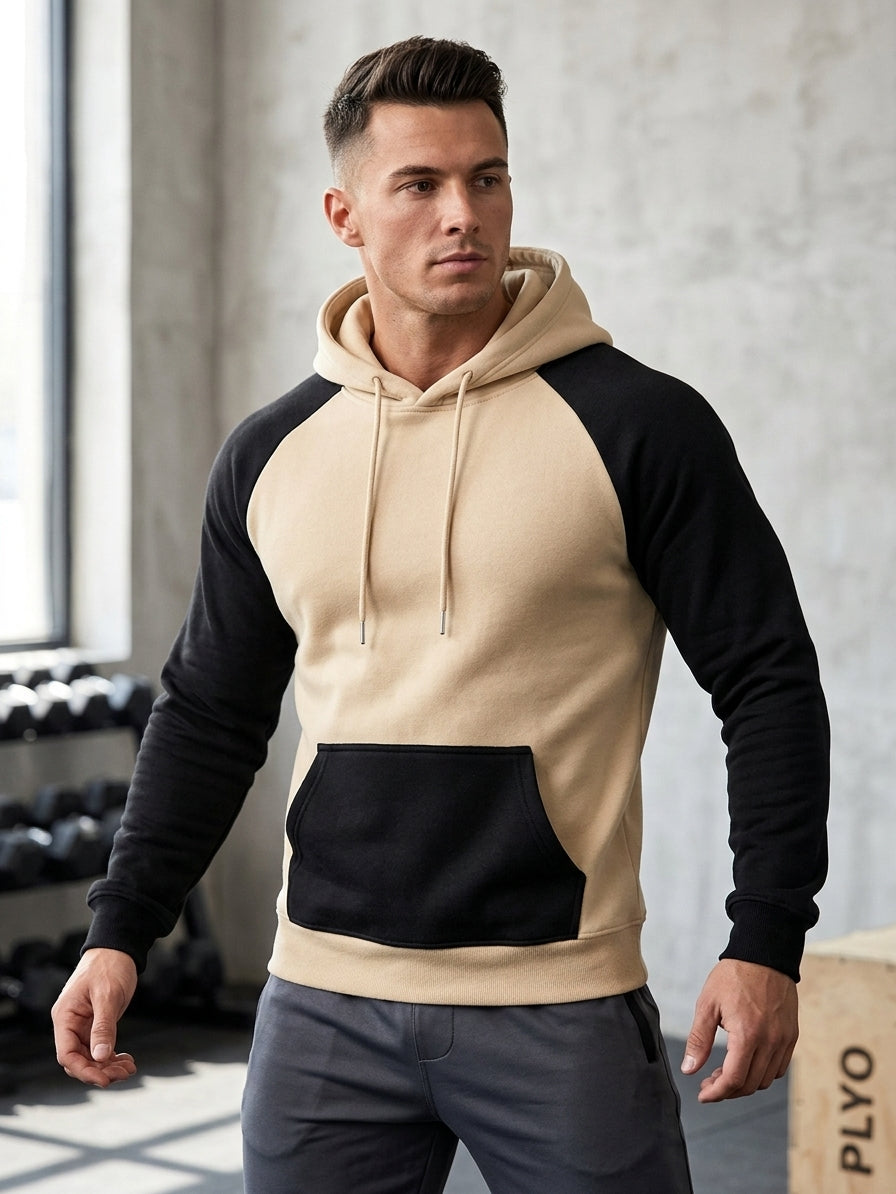 A man wearing a colorblock raglan hoodie with a beige body and sleeves, black shoulders and hood, and a black kangaroo pocket, paired with gray sweatpants in a gym setting.