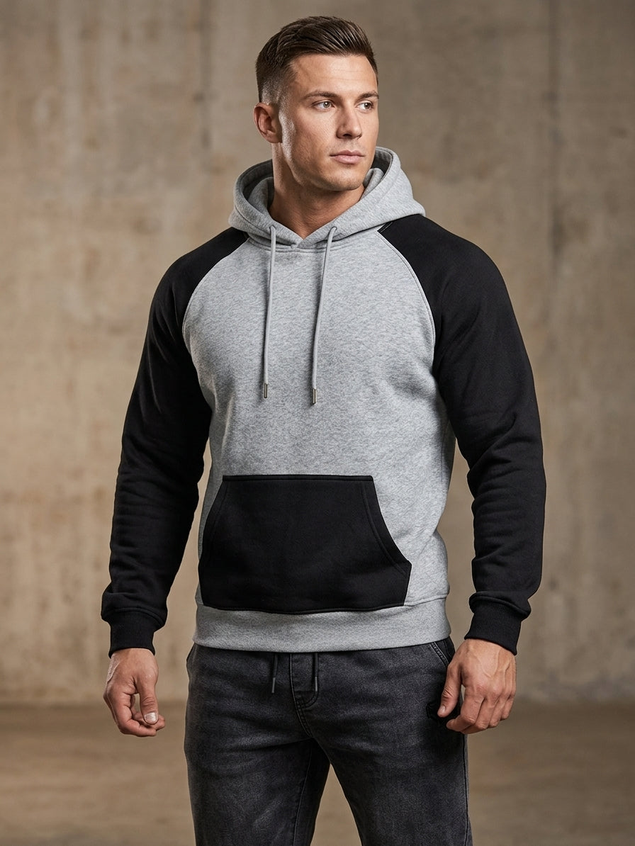 A man modeling a colorblock raglan hoodie with a heather gray body and black sleeves, hood, and front pocket, paired with dark gray jeans against a textured concrete wall.