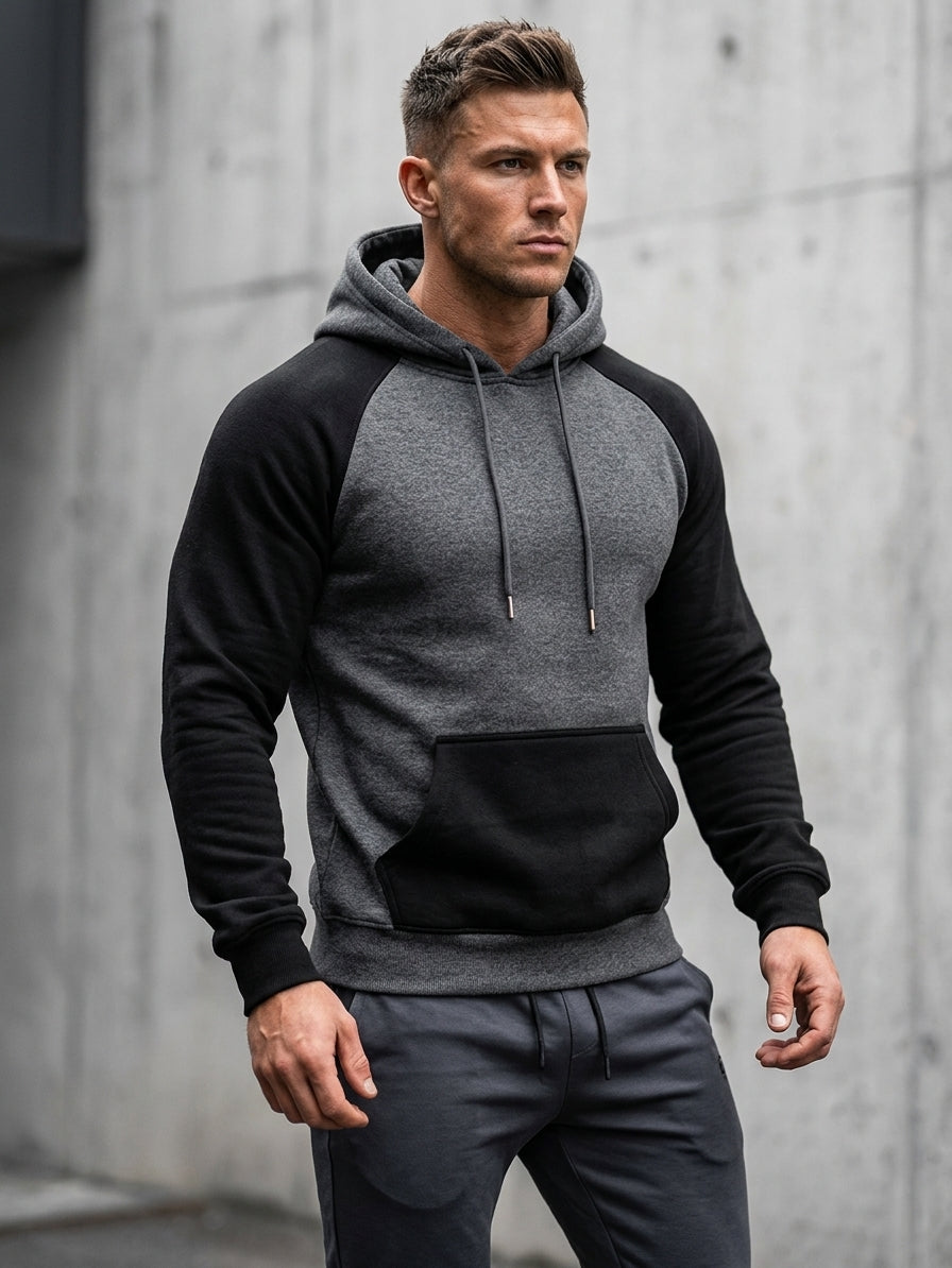 A man wearing a colorblock raglan hoodie with a heather gray body and sleeves, solid black shoulders, and a black kangaroo pocket, paired with dark gray sweatpants, standing against a concrete wall.
