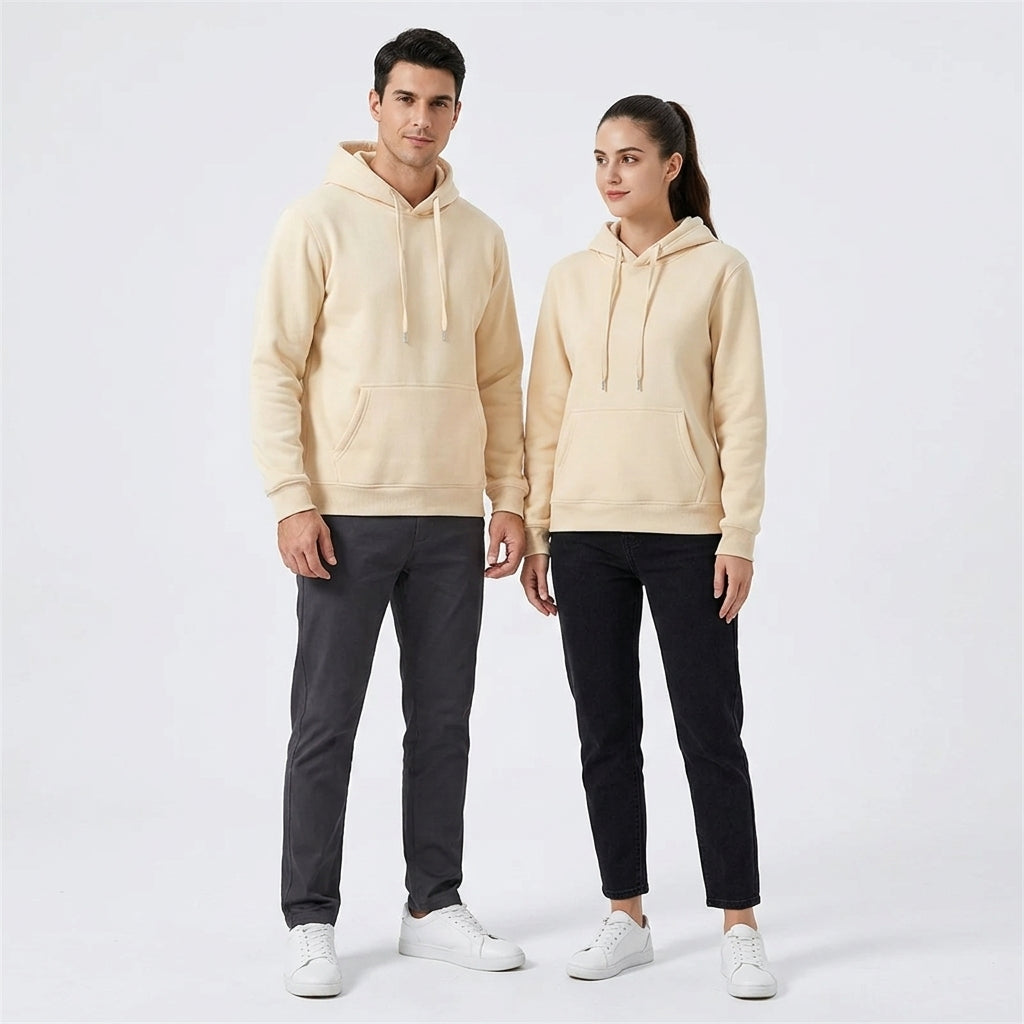 A man and a woman standing side by side in a studio, wearing matching light beige hooded sweatshirts with drawstrings and front kangaroo pockets, paired with dark pants and white sneakers.