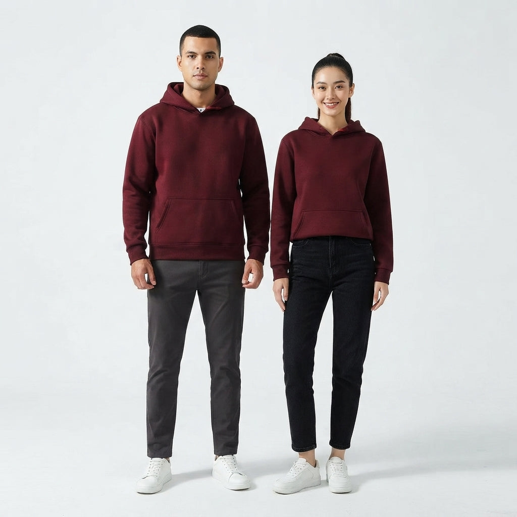 A man and a woman modeling matching burgundy pullover hoodies, standing against a clean light gray studio background. The man wears dark gray pants and white sneakers, while the woman wears black pants and white sneakers.