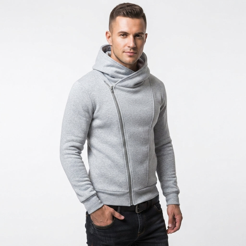 A man wearing a light gray zip-up hoodie with a high folded collar, paired with dark jeans, photographed against a plain light gray studio background.