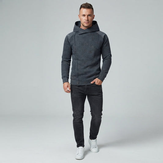 A man modeling a dark gray hooded sweatshirt with contrasting dark gray sleeves, paired with dark gray jeans and white sneakers, against a light gray studio background.