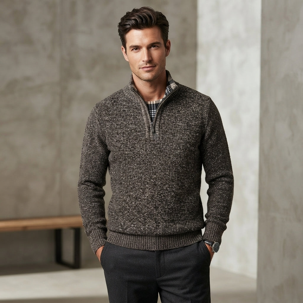 A man wearing a dark gray and brown heathered knit quarter-zip sweater over a plaid shirt and dark trousers, standing in a modern interior setting with textured gray walls.