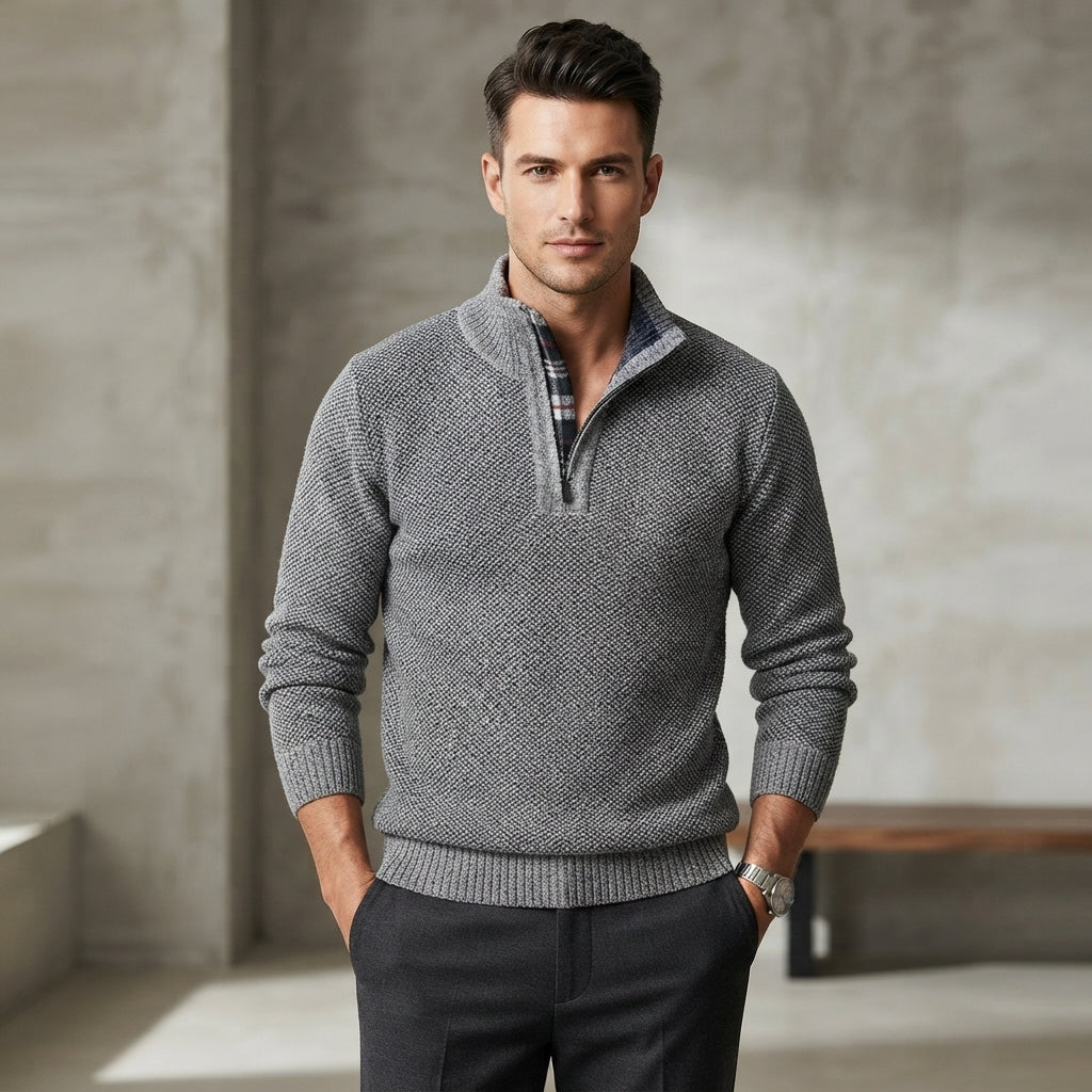 A man wearing a gray textured quarter-zip knit sweater over a plaid shirt and dark trousers, standing in a modern interior with a concrete wall background.