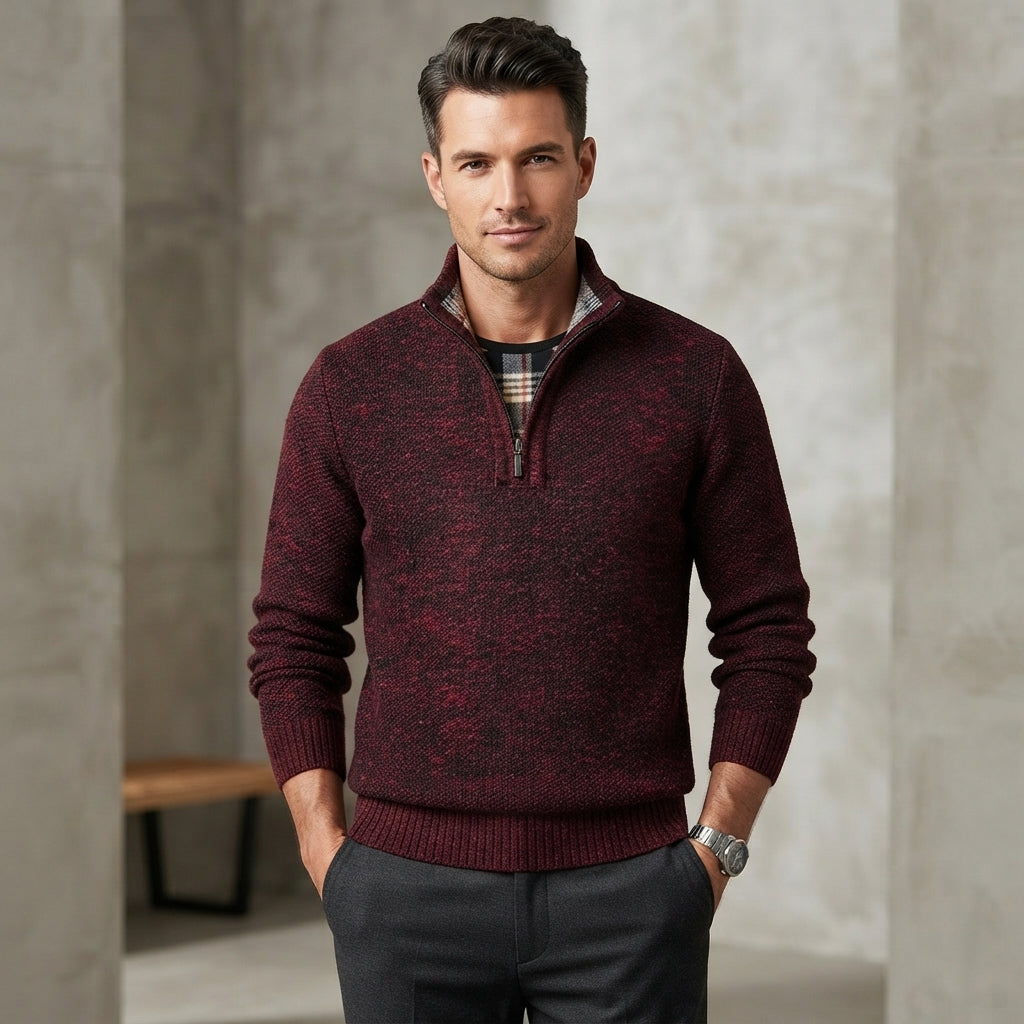 A man wearing a marled burgundy and black quarter-zip knit sweater over a plaid shirt, paired with dark gray trousers, standing in a modern indoor setting with textured gray walls.