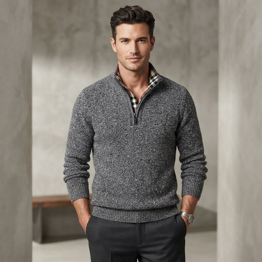 A man wearing a dark gray heathered quarter-zip knit sweater with a black and white plaid collar lining, paired with dark gray trousers, standing in a modern concrete hallway.
