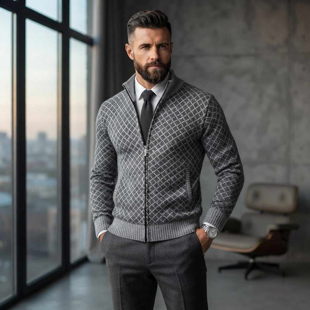 A man wearing a gray zip-up cardigan with a geometric diamond pattern over a white collared shirt and dark tie, standing in a modern room with large windows and a concrete wall.