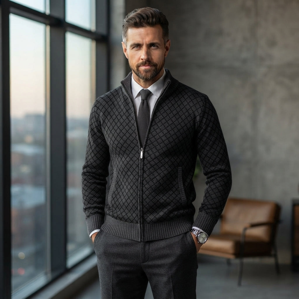 A man wearing a dark gray zip-up knit cardigan with a diamond pattern, styled over a white collared shirt and a dark gray tie, standing in a modern office setting with large windows and a leather chair in the background.