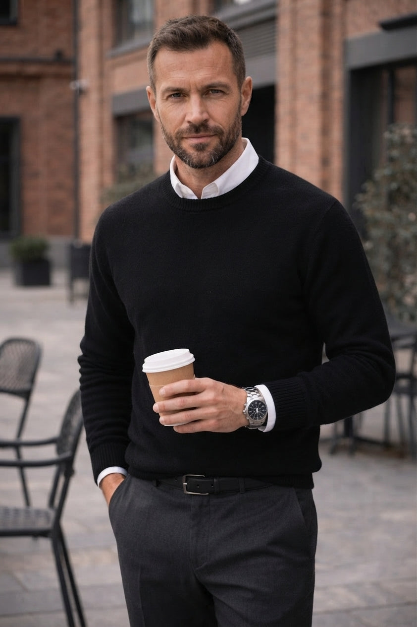 A man wearing a black crewneck sweater over a white collared shirt, holding a paper coffee cup in an outdoor urban setting.