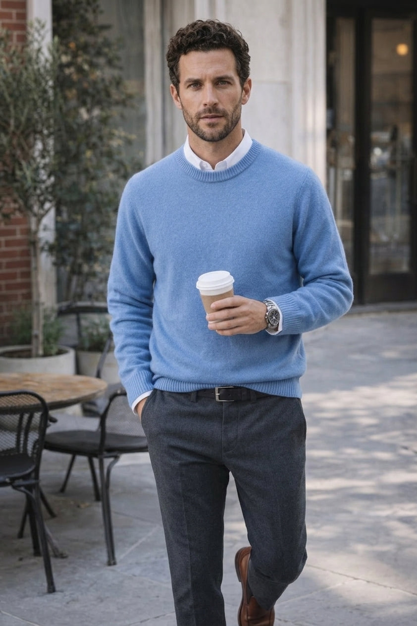 A man wearing a light blue crewneck sweater over a white collared shirt, paired with dark gray trousers and brown leather shoes, holding a coffee cup in an outdoor cafe setting.