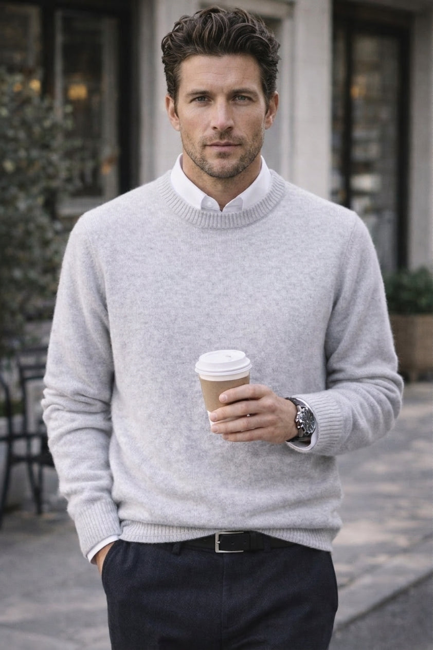 A man wearing a light gray knit crewneck sweater over a white collared shirt, paired with dark trousers and holding a paper coffee cup in an outdoor urban setting.