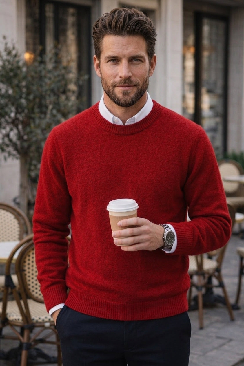 A man wearing a vibrant red knit crewneck sweater over a white collared shirt, standing on an outdoor cafe patio and holding a paper coffee cup.
