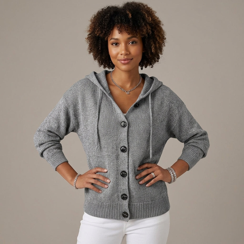 A woman with curly brown hair wearing a gray knit hooded cardigan with black buttons and white pants, standing against a solid taupe background.