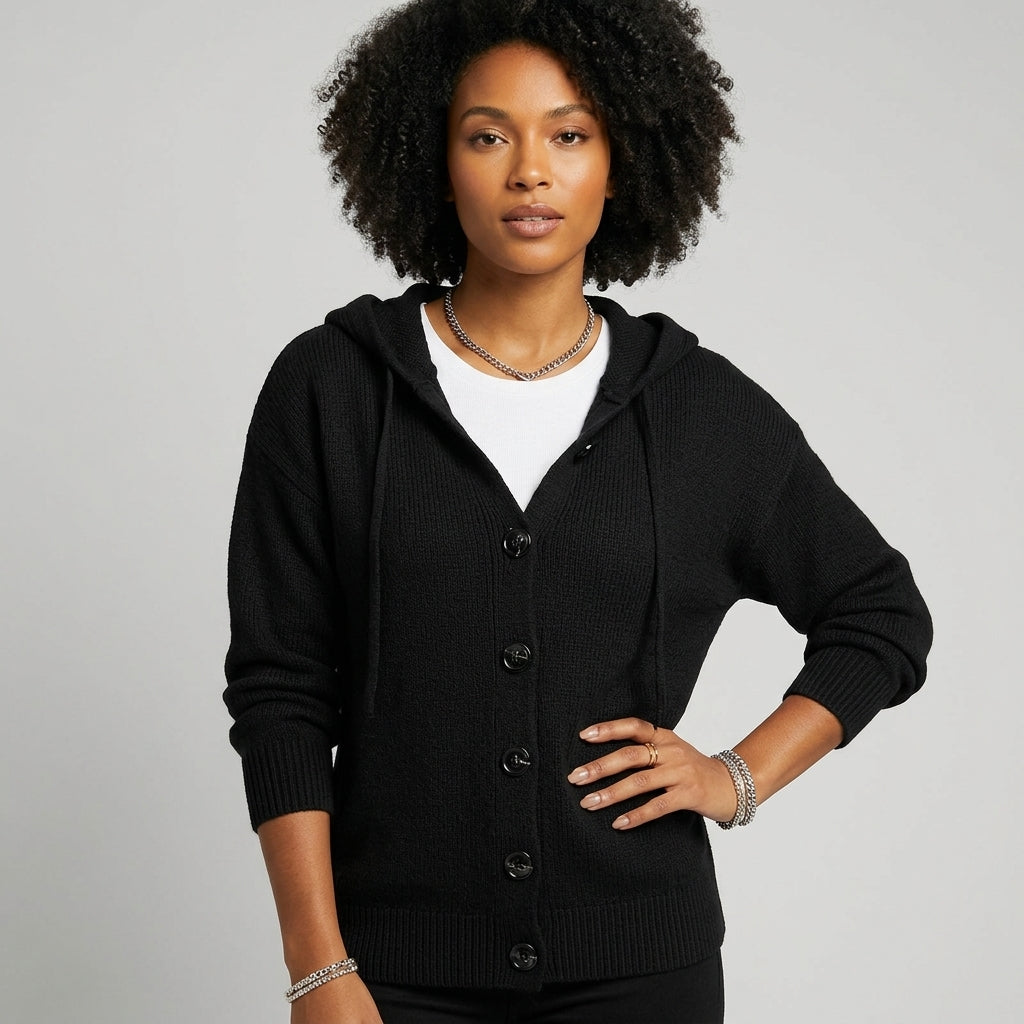 A woman with curly black hair wearing a black ribbed knit hooded cardigan over a white crewneck shirt, standing against a light gray studio background.