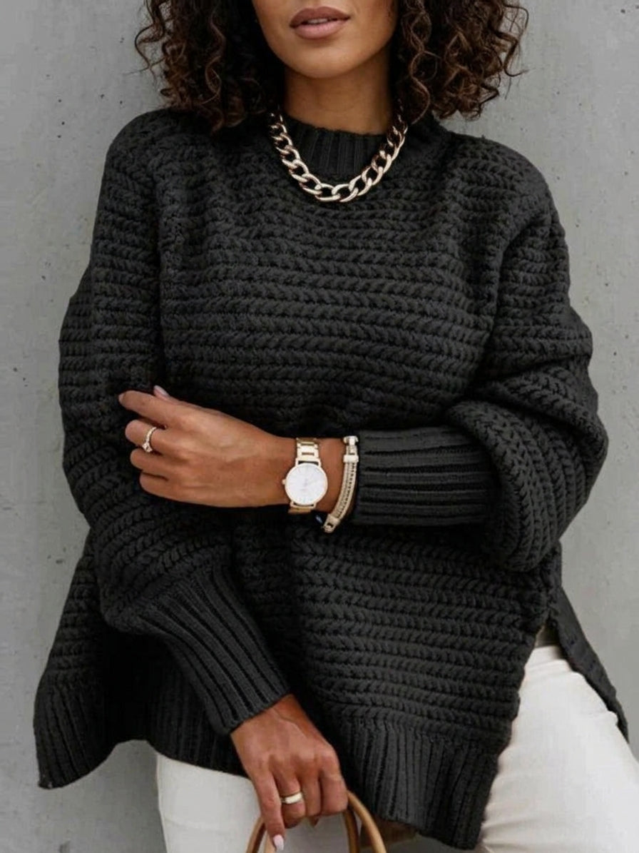 A woman wearing a dark charcoal gray chunky knit sweater, styled with a chunky gold chain necklace, a gold watch, and white pants, standing against a gray concrete wall.