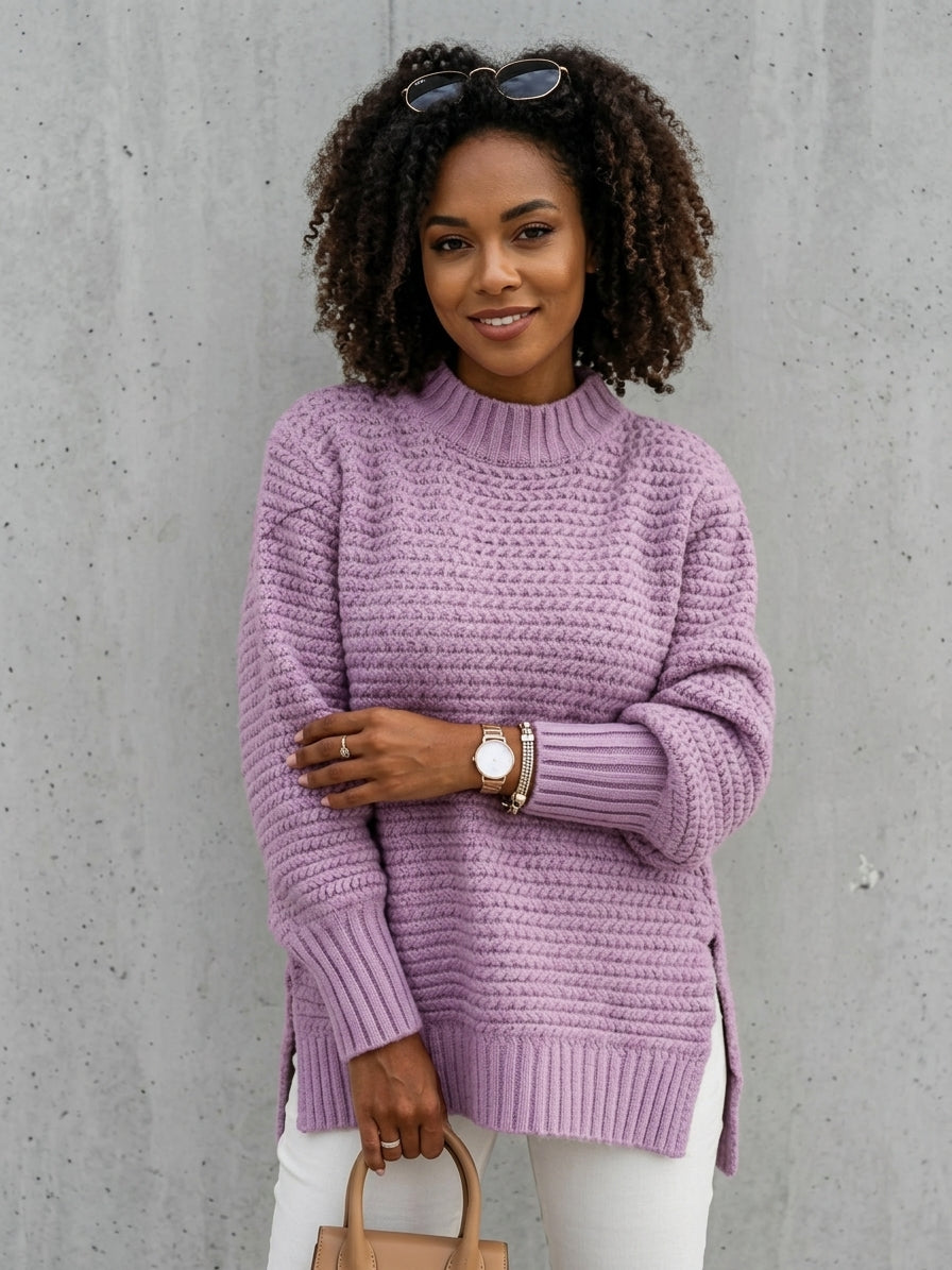 A woman wearing a chunky knit lavender-purple sweater and white pants, standing against a gray concrete wall and holding a tan handbag.