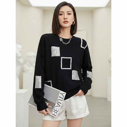 A woman modeling a black long-sleeve sweater with silver rhinestone square and rectangular patchwork details, paired with white shorts and holding a magazine in a bright, modern indoor setting.