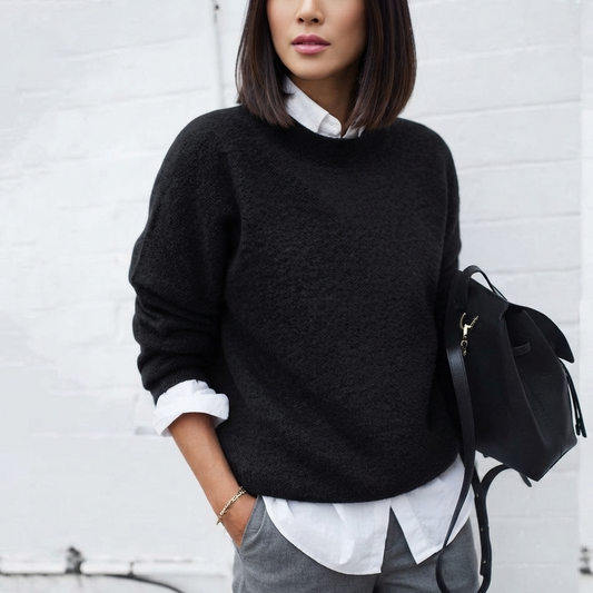 A woman wearing a black fuzzy knit sweater over a white collared shirt, paired with gray trousers and carrying a black backpack, standing against a white brick wall.