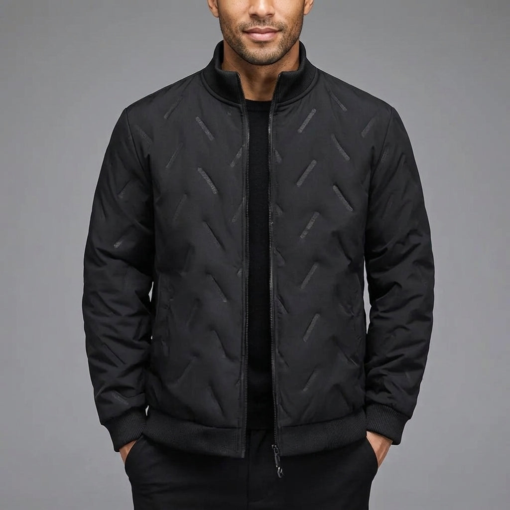 A man wearing a black quilted bomber jacket with a subtle geometric pattern, paired with a black shirt and trousers, set against a solid gray studio background.