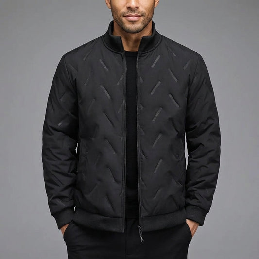 A man wearing a black quilted bomber jacket with a subtle geometric pattern, paired with a black shirt and trousers, set against a solid gray studio background.