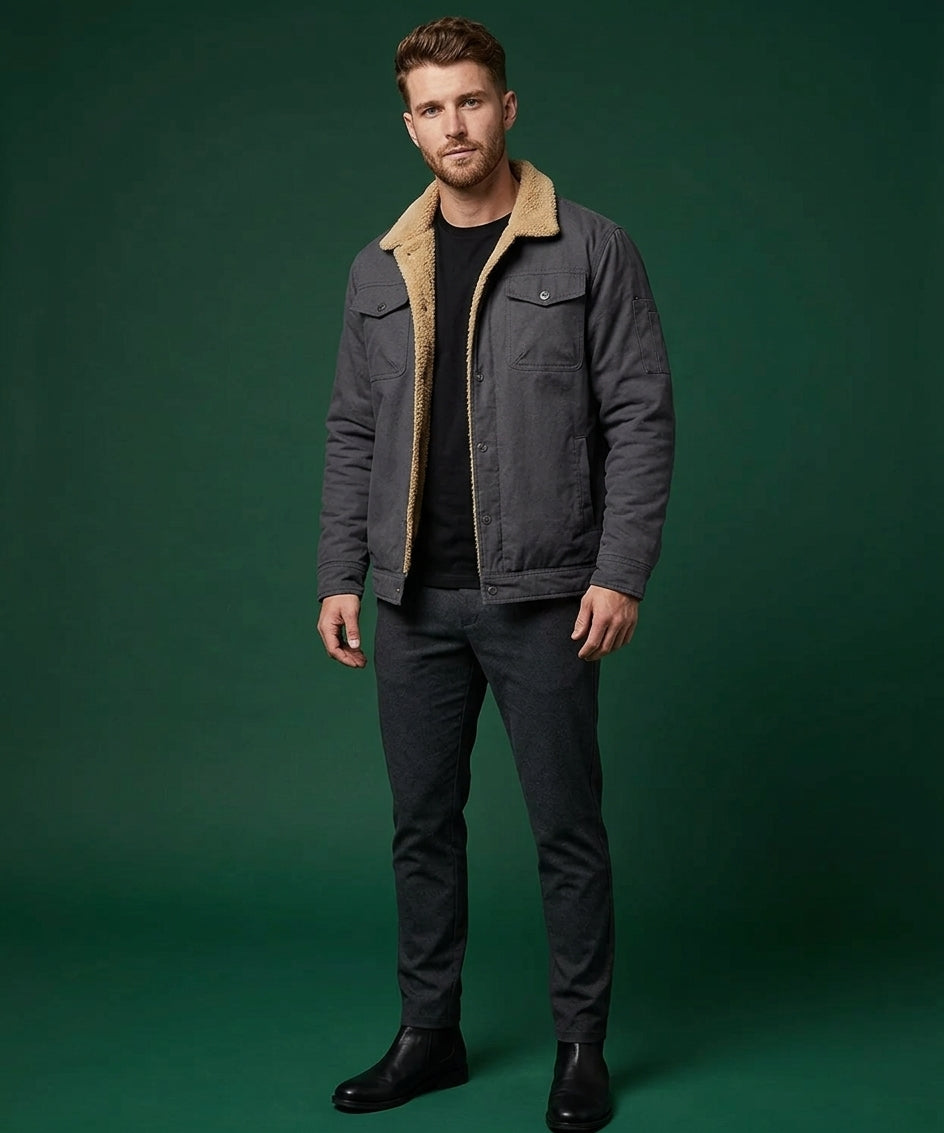 A man wearing a dark gray jacket with a tan sherpa collar, a black t-shirt, dark gray jeans, and black boots, standing against a solid dark green studio background.
